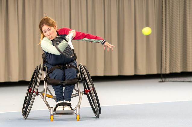 Sportlerin im Rollstuhl schl&auml;gt einen Tennisschlag mit einem Schl&auml;ger auf einem Indoor-Tennisplatz.
