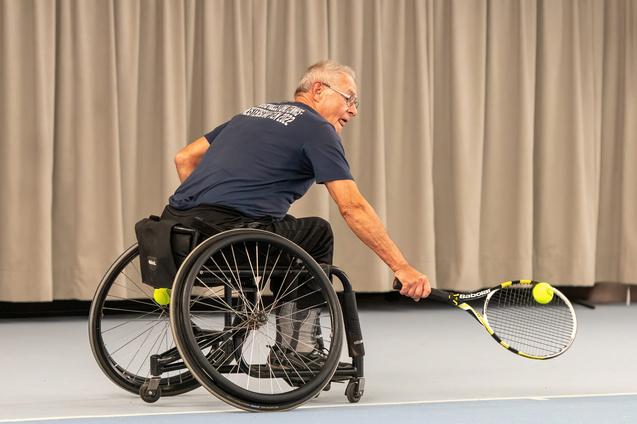 Mann im Rollstuhl schl&auml;gt mit einem Tennisracket einen Ball auf einem Tennisplatz mit grauer R&uuml;ckwand.