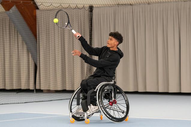 Junger Mann im Rollstuhl schl&auml;gt mit einem Tennisschl&auml;ger einen Ball auf einem Indoor-Tennisplatz.