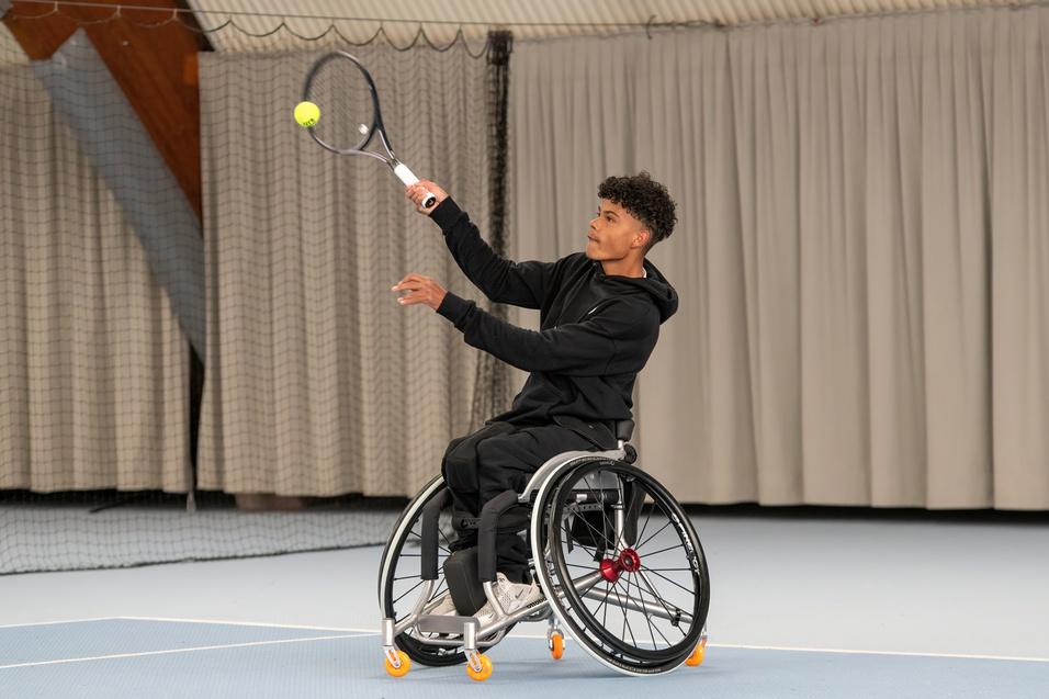 Junger Mann im Rollstuhl schl&auml;gt mit einem Tennisschl&auml;ger einen Ball auf einem Indoor-Tennisplatz.