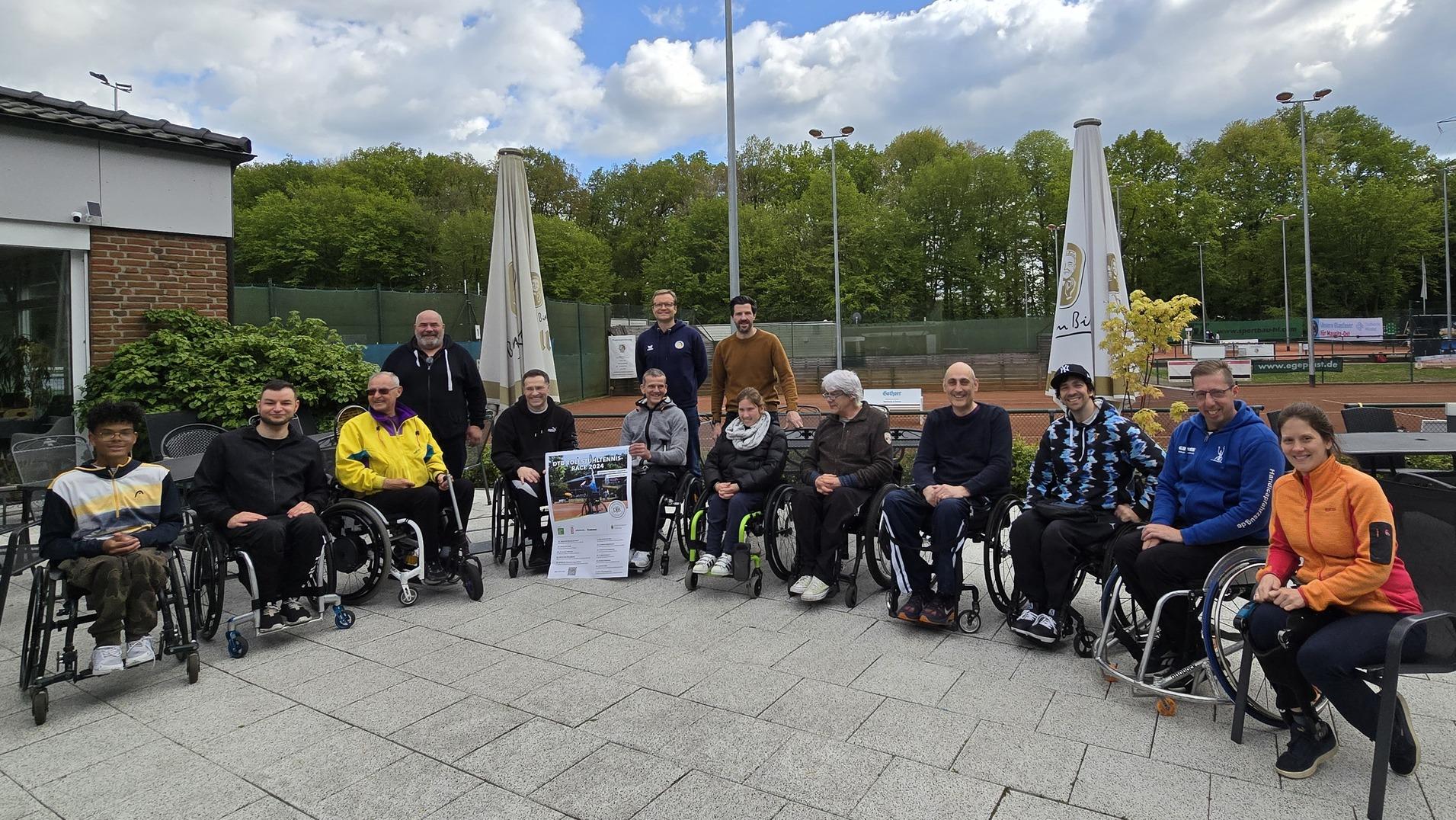 Gruppenaufnahme von 15 Personen im Rollstuhl, die vor einem Sportgel&auml;nde auf einer Terrasse sitzen.