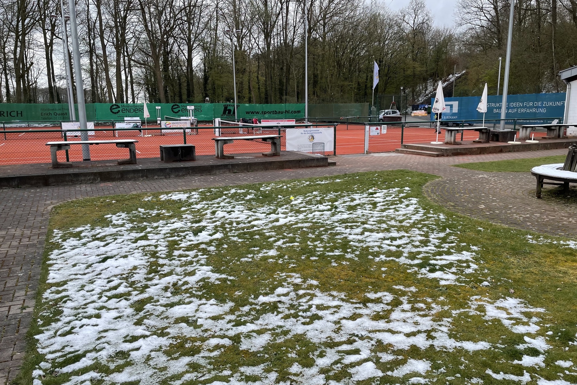 Tennisplatz mit orangefarbener Fl&auml;che und teils schneebedecktem, gr&uuml;nem Gras, umgeben von B&auml;umen und Geb&auml;uden.
