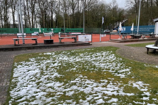 Tennisplatz mit orangefarbener Fl&auml;che und teils schneebedecktem, gr&uuml;nem Gras, umgeben von B&auml;umen und Geb&auml;uden.