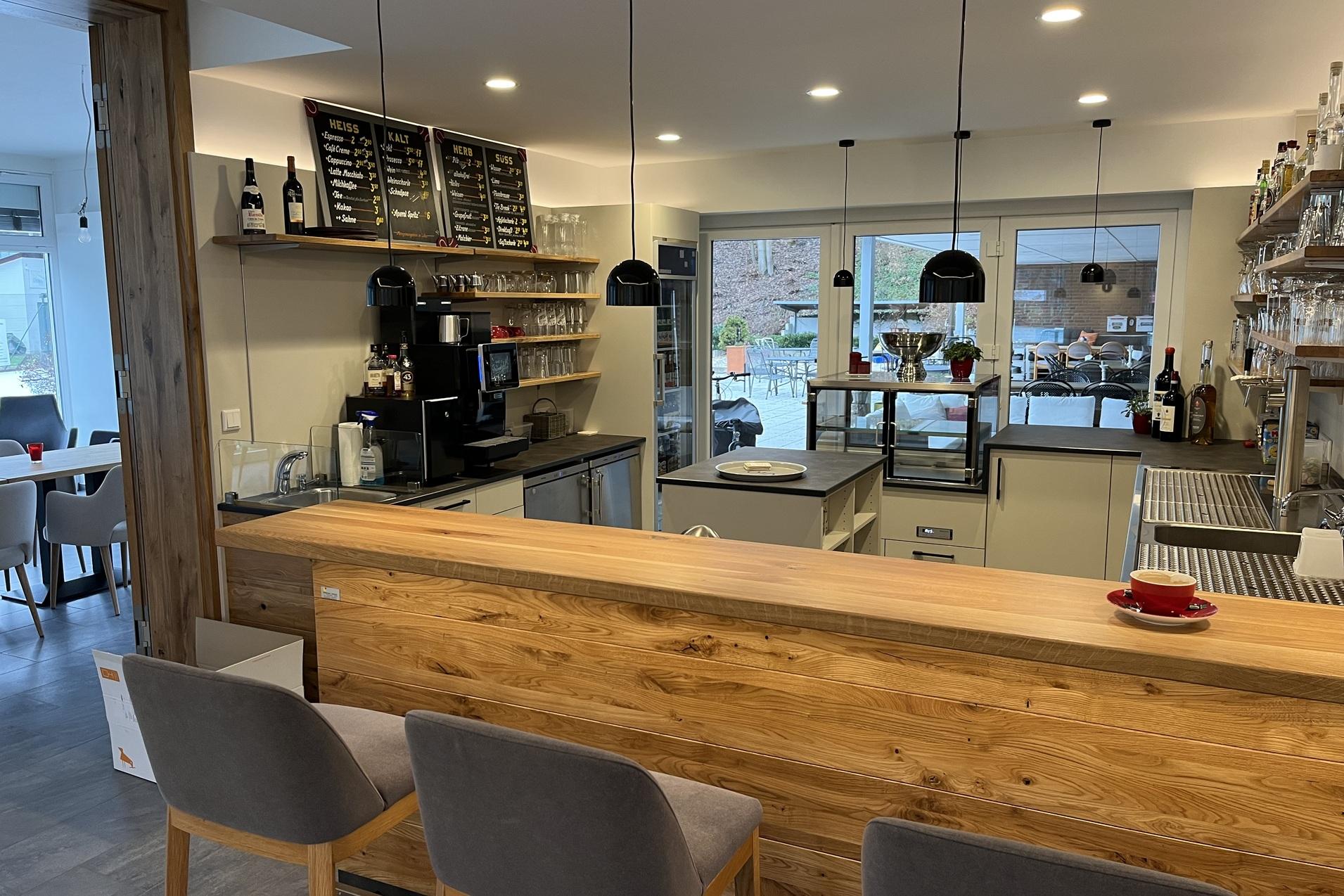 Moderne Caf&eacute;bar mit Holztheke, grauen St&uuml;hlen und offenem K&uuml;chenbereich. Tageslicht f&auml;llt durch gro&szlig;e Fenster.