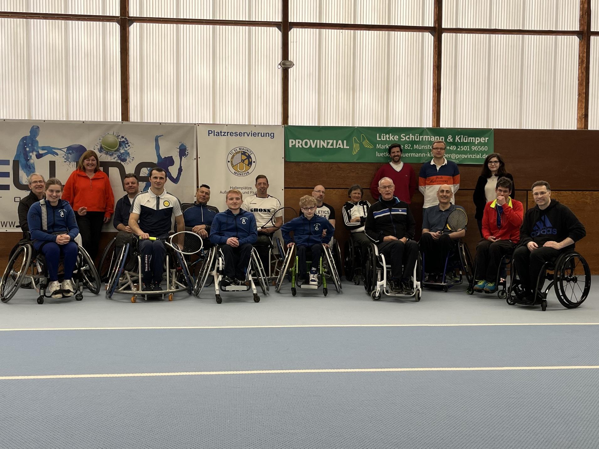 Gruppenfoto von 16 Personen im Rollstuhl auf einem Sportplatz, umgeben von Bannern und Sponsorenlogos.