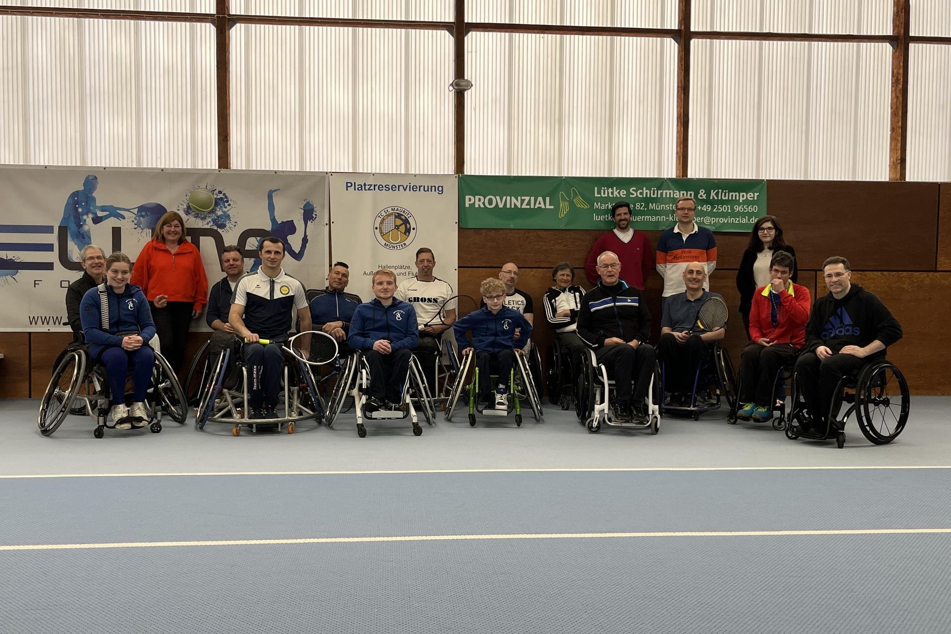 Gruppenfoto von 16 Personen im Rollstuhl auf einem Sportplatz, umgeben von Bannern und Sponsorenlogos.