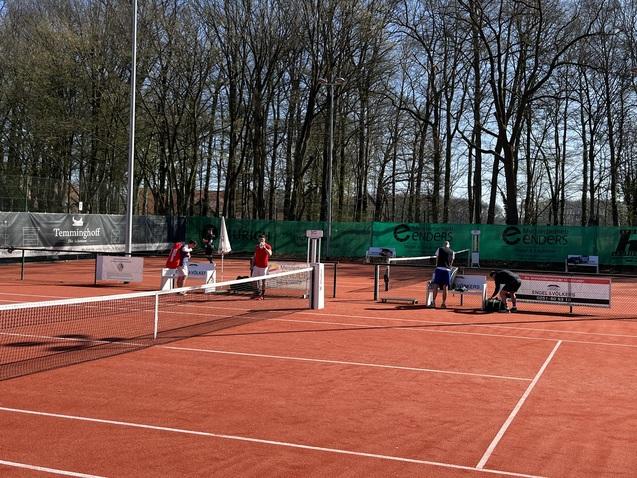 Tennisplatz mit mehreren Spielern in Aktion und B&auml;ume im Hintergrund, bei sonnigem Wetter auf rotem Sand.