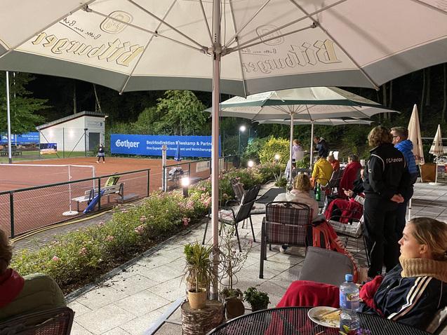 Auf einer Terrasse sitzen Zuschauer unter Schirmen und beobachten ein Tennisspiel auf einem beleuchteten Platz.