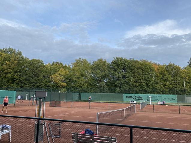 Tennisplatz mit zwei Spielern beim Spiel, umgeben von gr&uuml;nen B&auml;umen und bew&ouml;lktem Himmel. Sitzbank im Vordergrund.