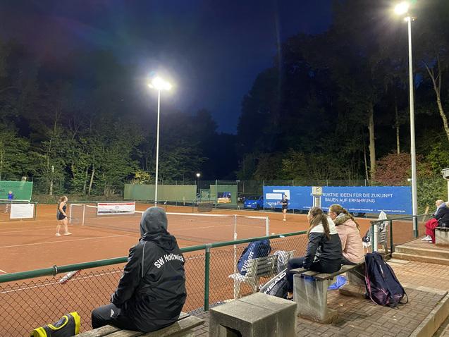 Tennisplatz bei Nacht mit einer Spielerin im Hintergrund und Zuschauern auf einer Bank im Vordergrund.