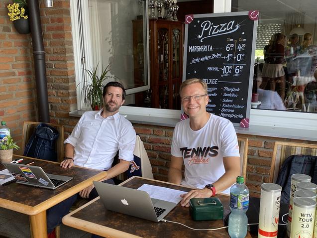 Zwei M&auml;nner sitzen an Tischen im Freien, arbeiten an Laptops und haben Getr&auml;nke vor sich. Im Hintergrund steht eine Speisekarte.
