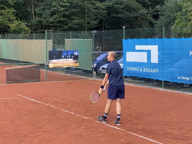 Tennis-Spieler auf einem roten Platz, bereit zum Aufschlag, umgeben von gr&uuml;nen B&auml;umen und einem Werbebanner.