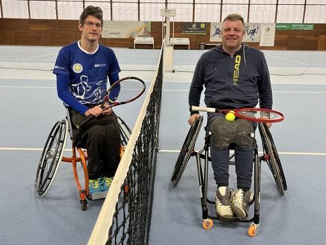 Zwei Rollstuhltennisspieler mit Schl&auml;gern und einem Ball sitzen auf gegen&uuml;berliegenden Seiten des Netzes in einem Tennisplatz.