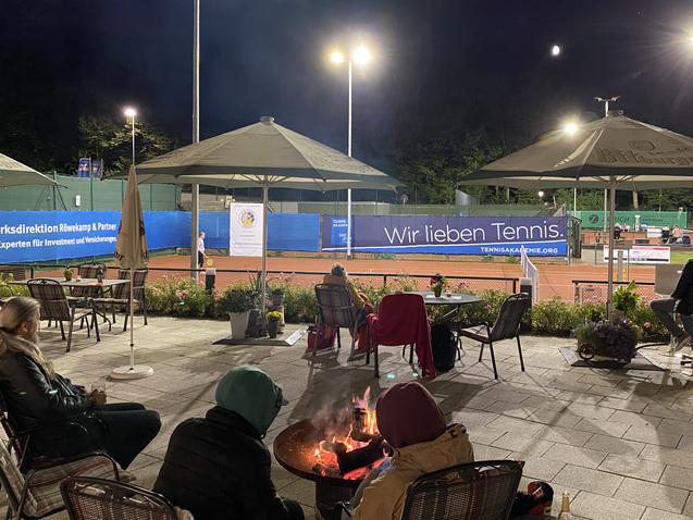 Tennisplatz bei Nacht mit Spielern, &uuml;berdachten Sitzbereichen und einer Feuerschale im Vordergrund. Beleuchtung und Banner sichtbar.