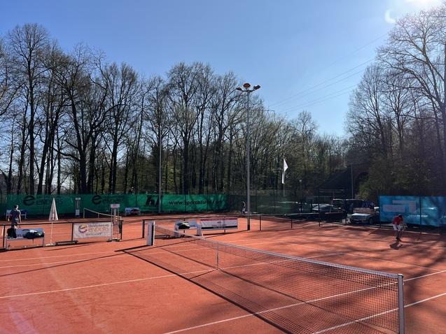 Tennisplatz mit rotem Sand, B&auml;umen im Hintergrund und Spielern bei sonnigem Wetter.