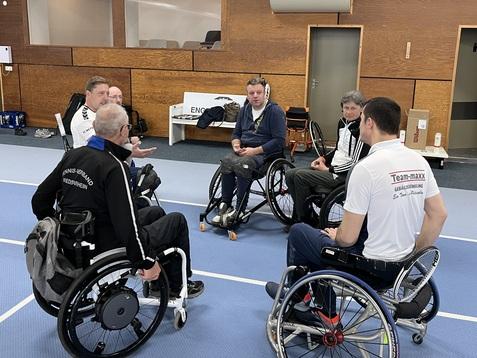 Sechs Rollstuhlfahrer sitzen in einem Sportzentrum und diskutieren auf einer blauen Sportfl&auml;che.