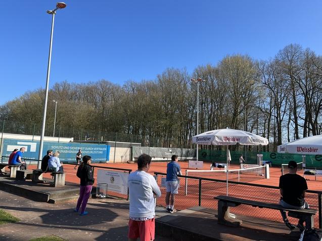 Gruppierung von Personen auf B&auml;nken an einem Tennisplatz, im Hintergrund Tennis-Spieler unter blauem Himmel.