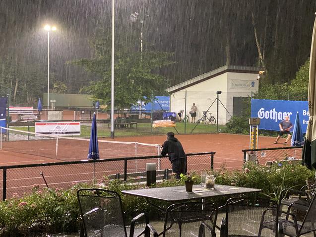 Regnerische Szenerie mit einem Tennisspieler auf einem roten Platz, Blick von einem &uuml;berdachten Sitzbereich.