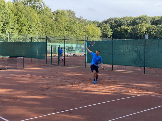 Tennisspieler in blauem Shirt schl&auml;gt einen Ball auf einem Sandplatz mit gr&uuml;nen Umz&auml;unungen und B&auml;umen im Hintergrund.