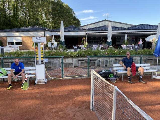 Zwei M&auml;nner sitzen auf B&auml;nken neben einem Tennisplatz, der Hintergrund zeigt ein Clubhaus und B&auml;ume.