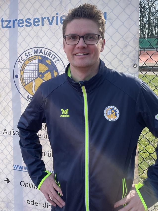 Mann mit Brille und Sportjacke steht l&auml;chelnd vor einem Banner mit dem Logo des TC St. Mauritz und Platzreservierungsinformationen.
