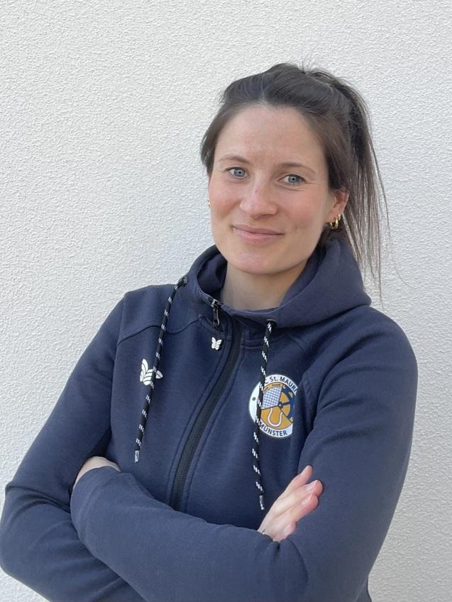 Portr&auml;t einer l&auml;chelnden Frau mit schulterlangen Haaren, die eng verschr&auml;nkte Arme vor einem neutralen Hintergrund h&auml;lt.