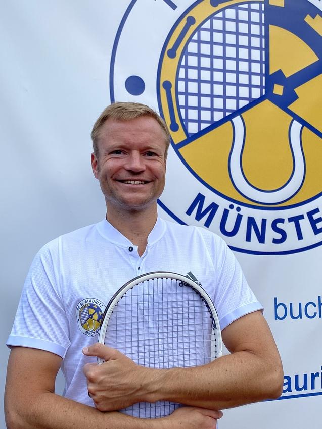 L&auml;chelnder Mann in wei&szlig;em Shirt h&auml;lt einen Tennisschl&auml;ger vor einem Banner des TC St. Mauritz M&uuml;nster.